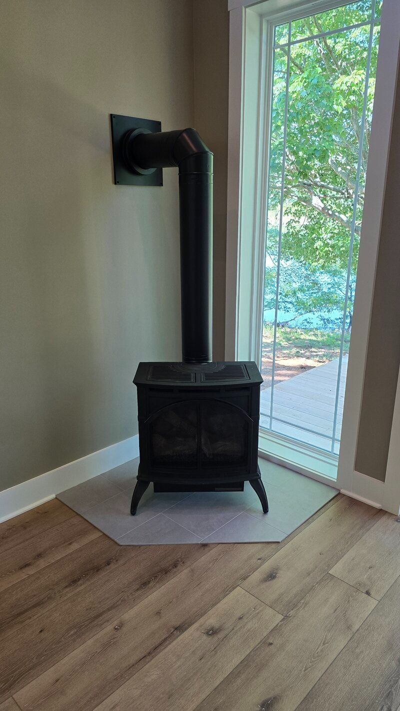 Black wood-burning stove with black chimney, in corner near a window overlooking trees.
