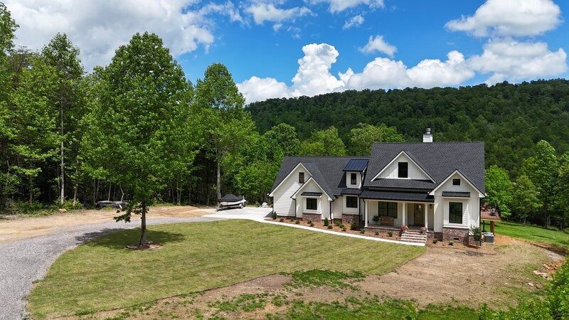 White house with black roof, nestled in green trees against a mountain backdrop under a blue sky.