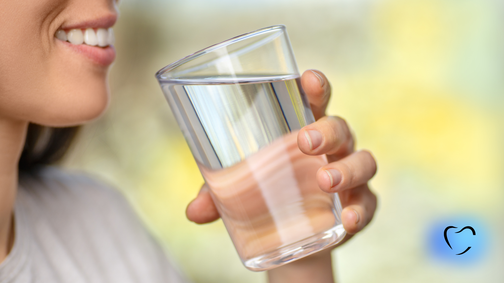 Woman drinking water from a glass; smiling; indoors, soft-focus background.