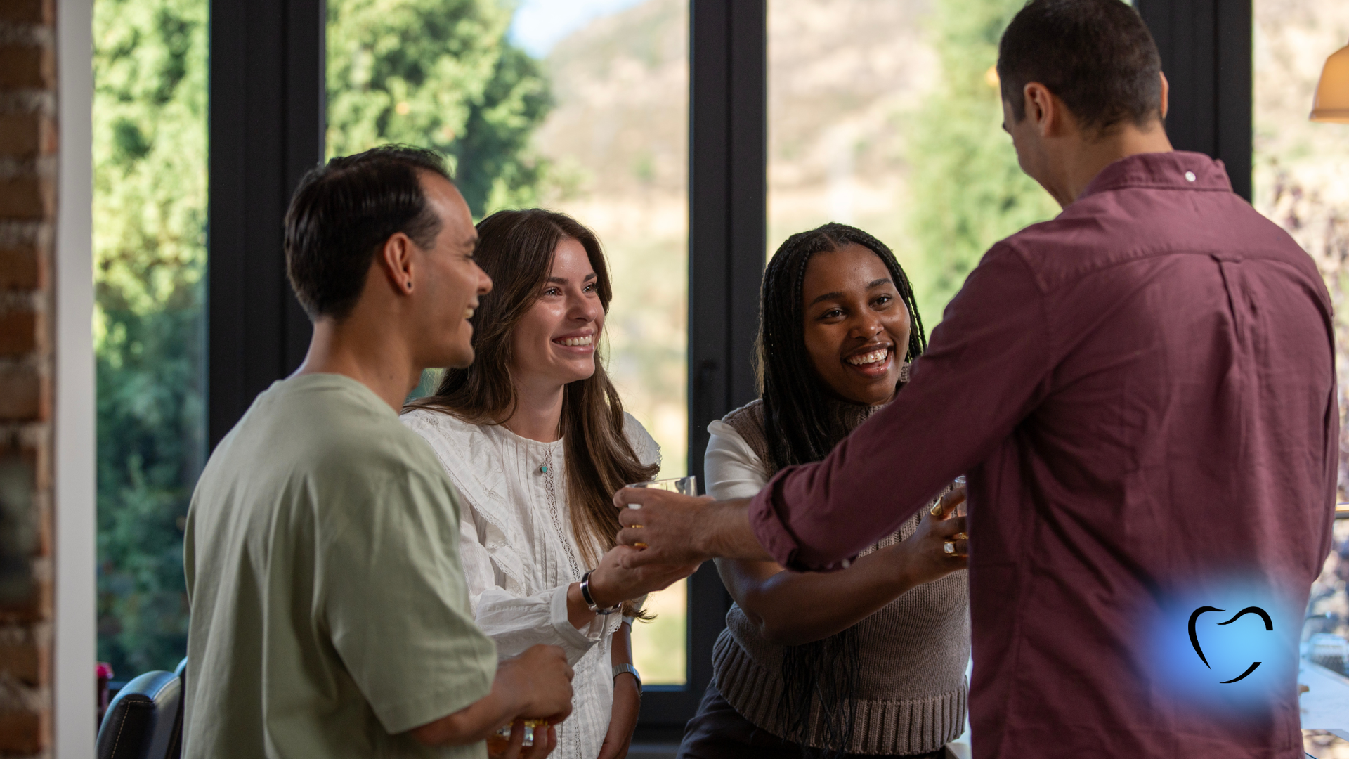 Four people laughing, greeting each other indoors. A sunny window is visible in the background.