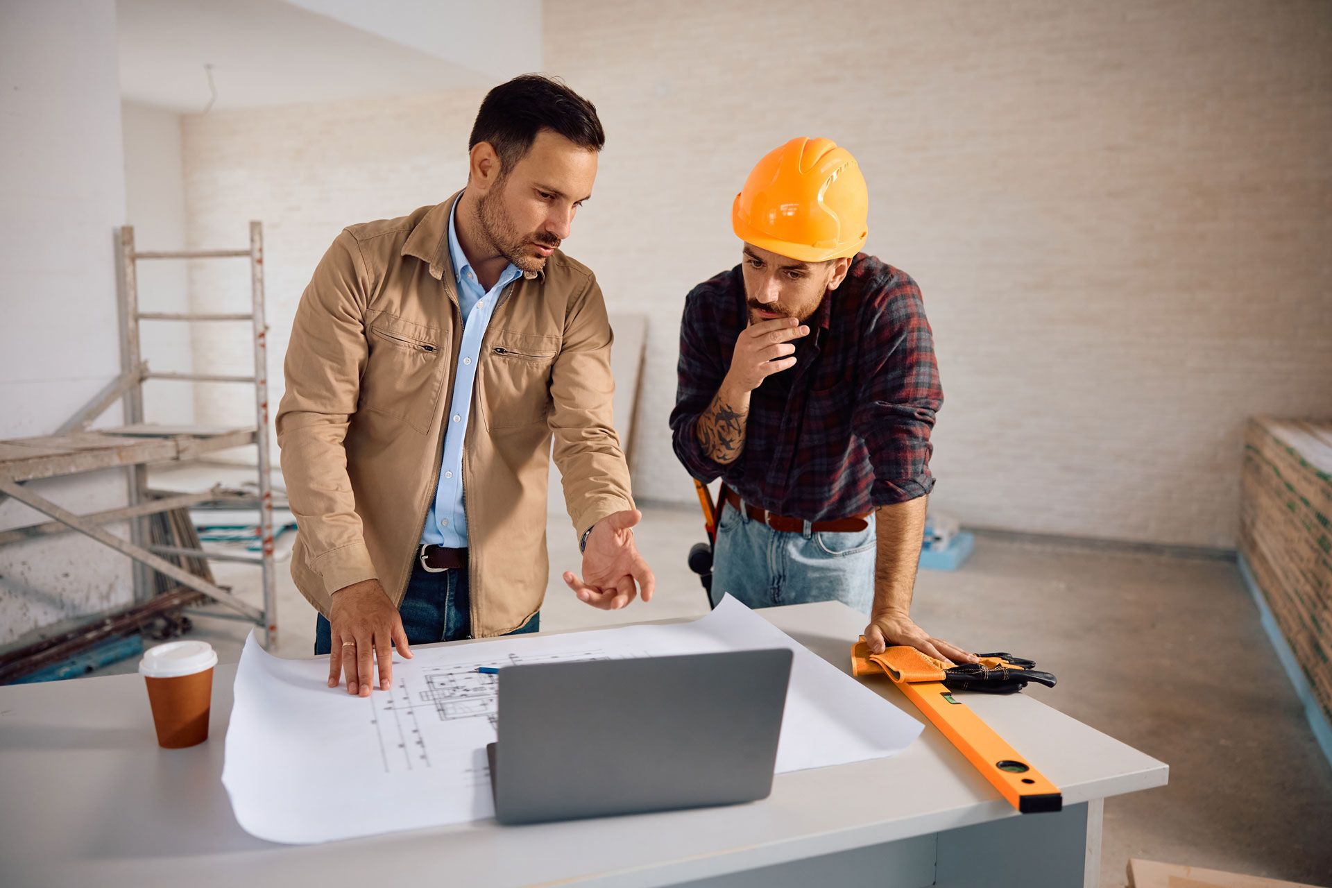 Two construction workers review blueprints and a laptop at a building site.