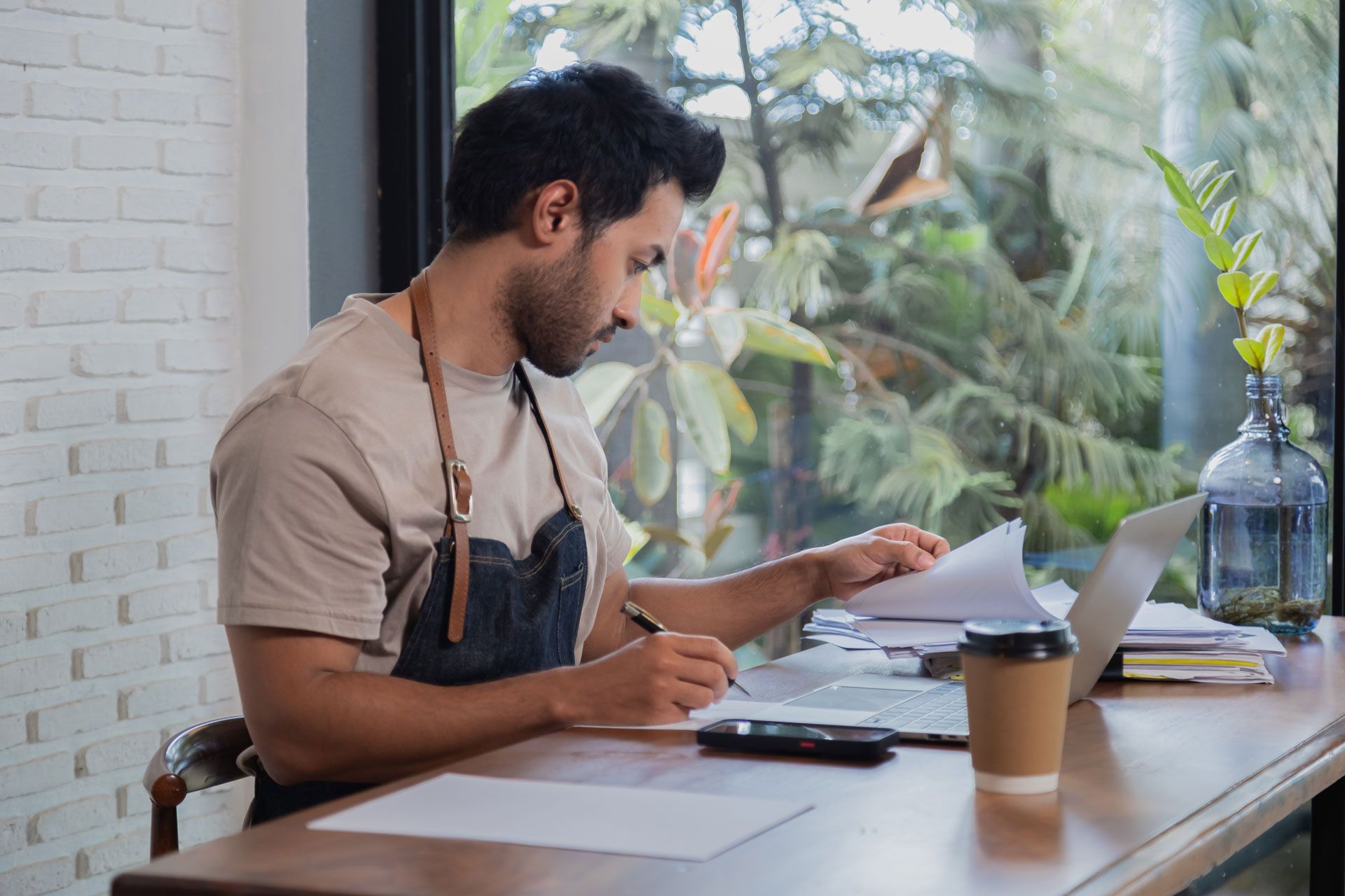 Man with apron at desk, writing on documents, working on laptop near window with foliage and coffee.