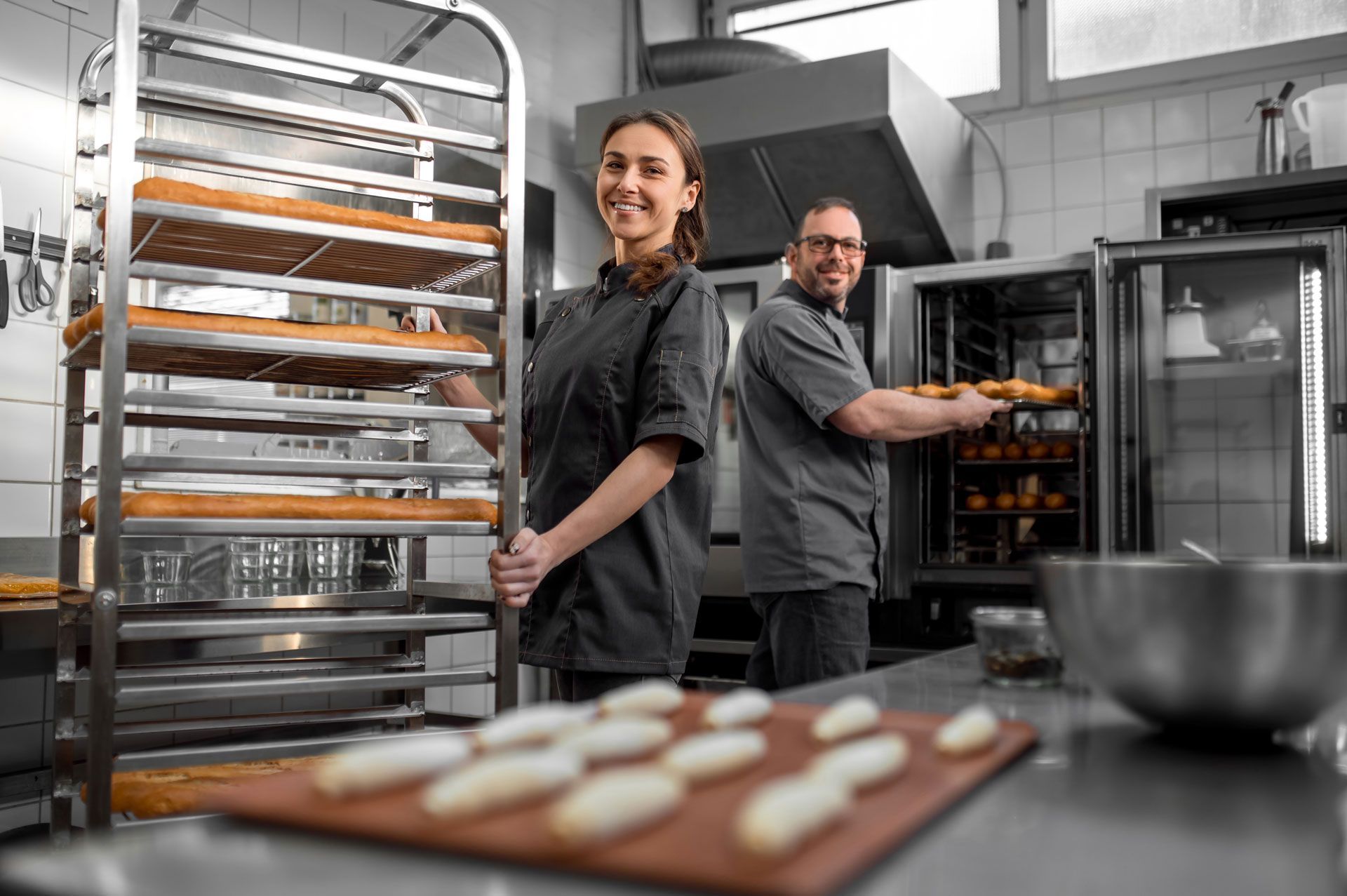Two bakers in a commercial kitchen; one smiles by a rack of baked goods, the other pulls loaves from an oven.