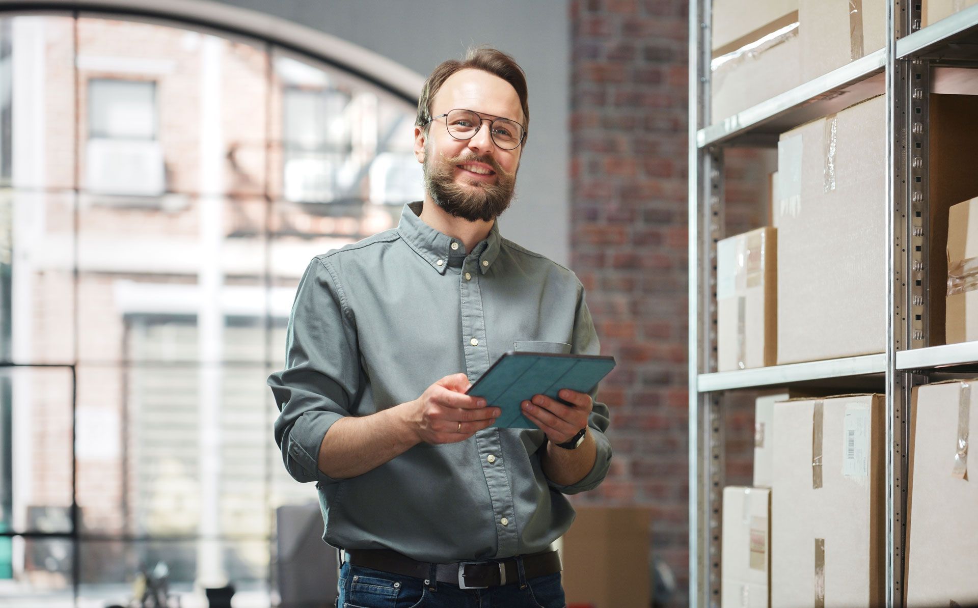 Man holding tablet, smiles in a warehouse. Boxes on shelves in the background.