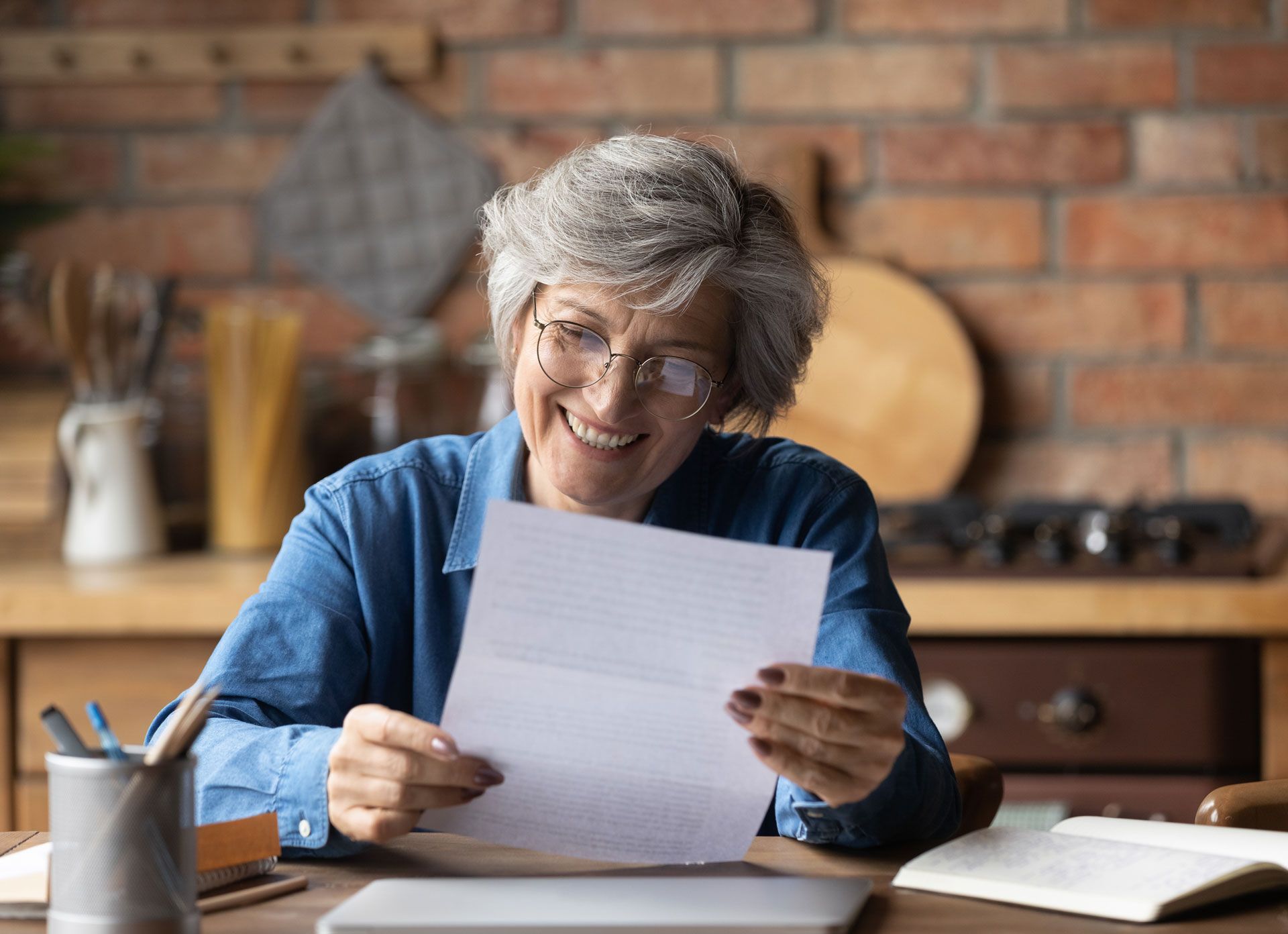 Woman with glasses smiles while reading a letter at a desk in a kitchen.