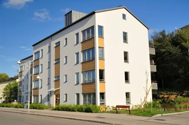 Modern apartment building with white and beige фасade, balconies, and greenery along a street