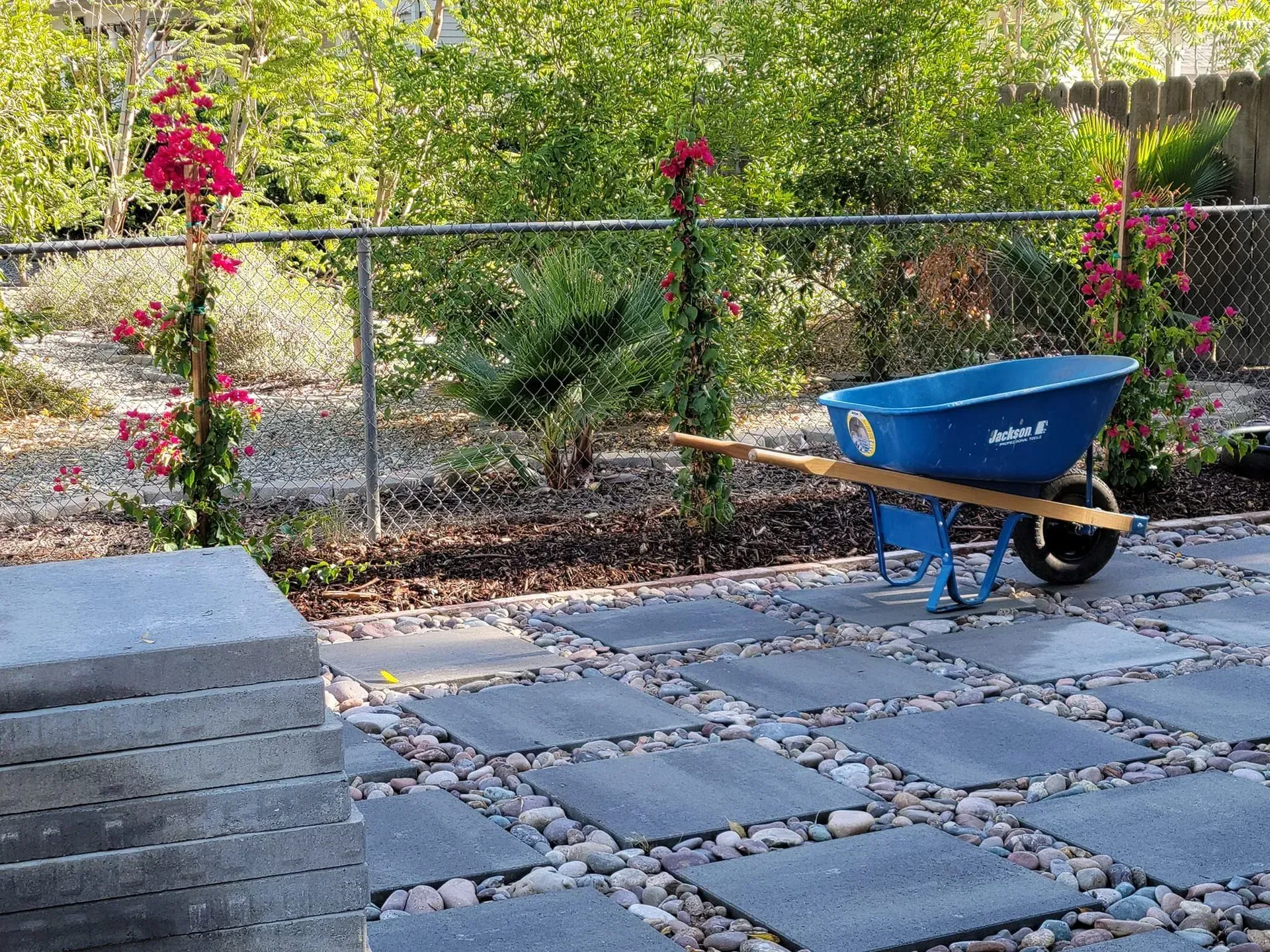 A blue wheelbarrow is parked on a patio next to a fence.