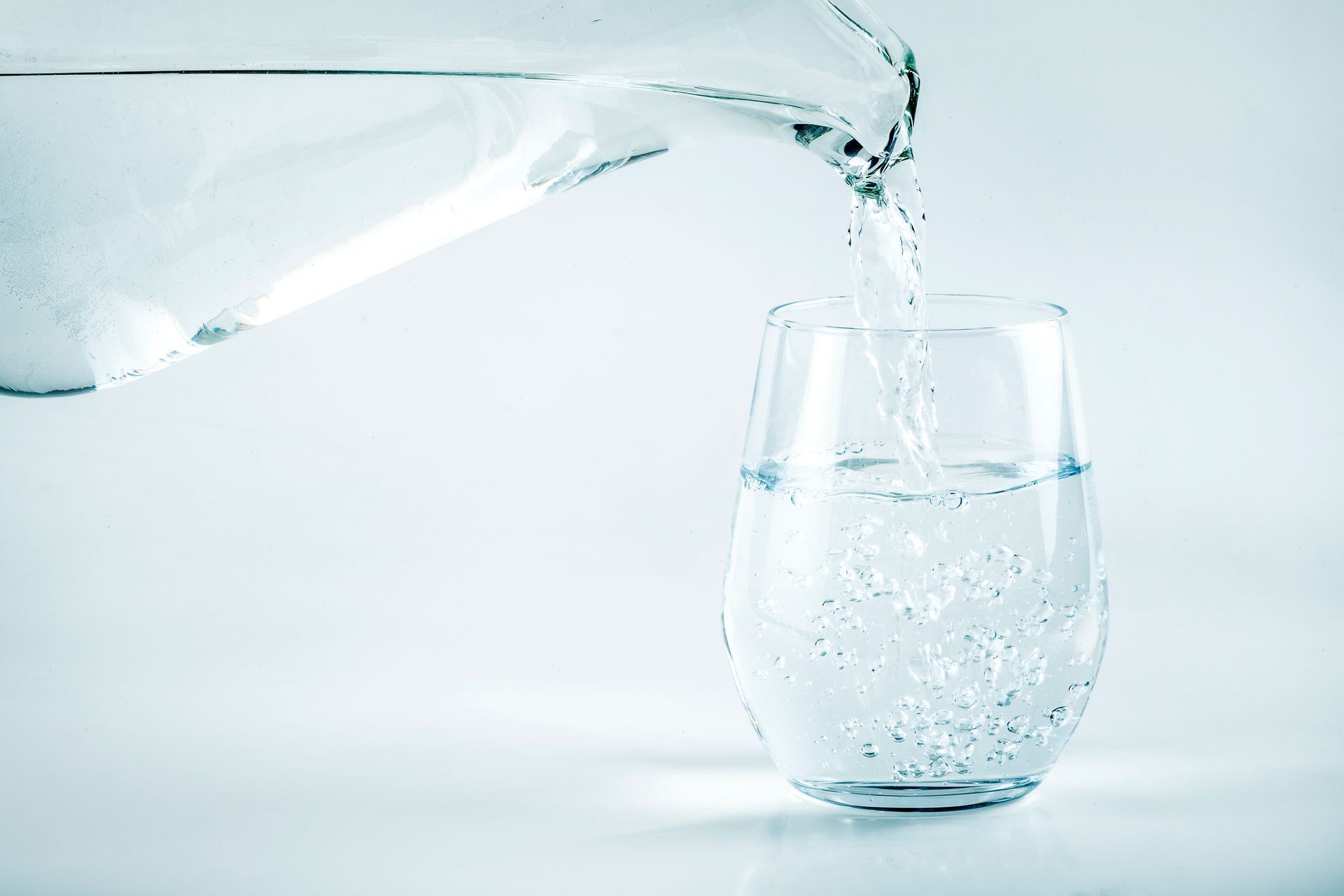 Water being poured from a pitcher into a glass, both filled with clear liquid.