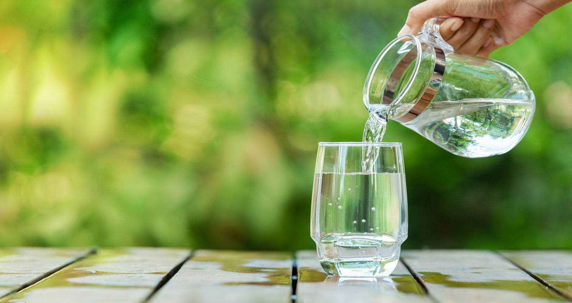 A hand pouring water from a pitcher into a glass on a wooden table, blurred green background.