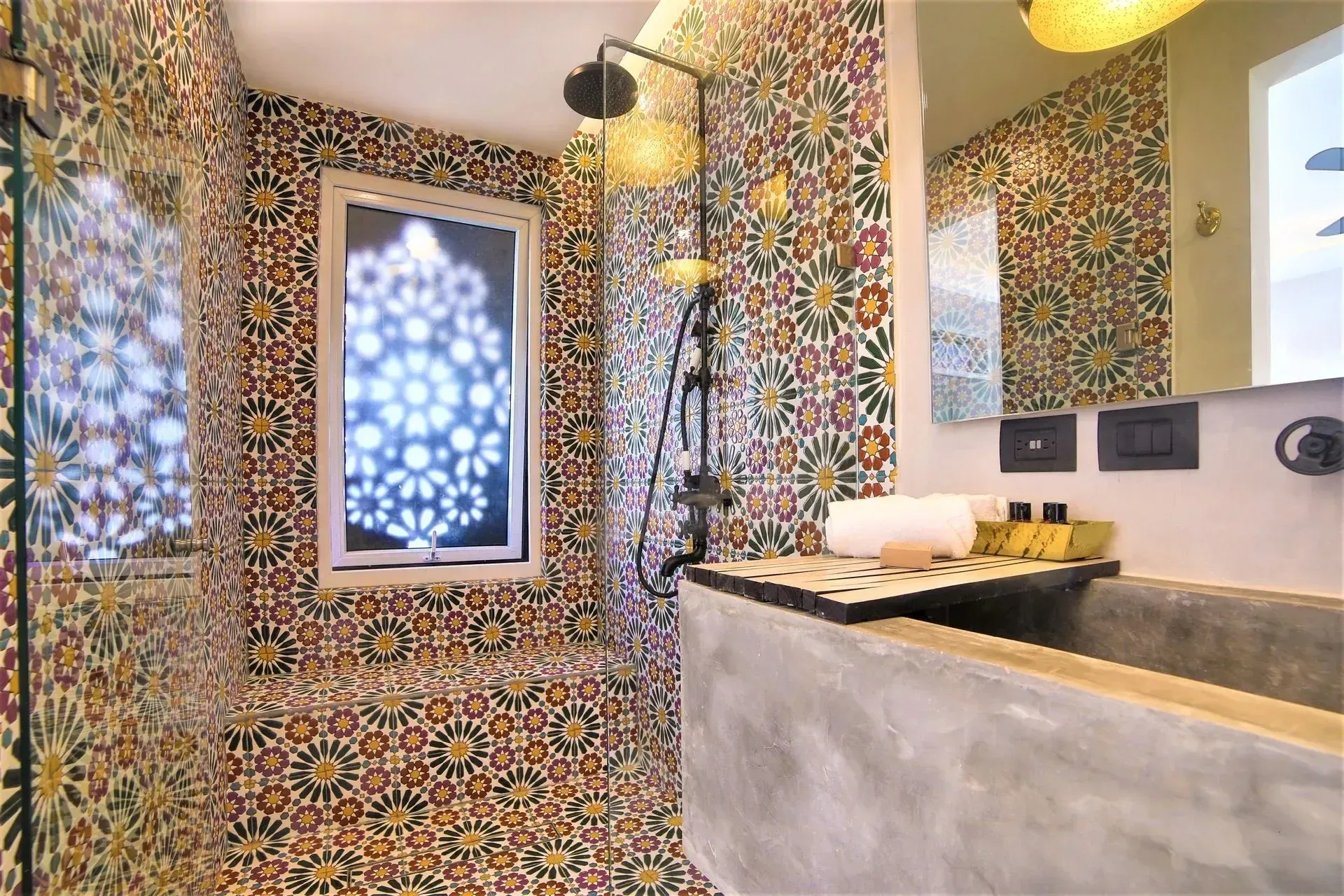 Bathroom with floral tile walls, concrete sink, window with decorative design, showerhead, and mirror.
