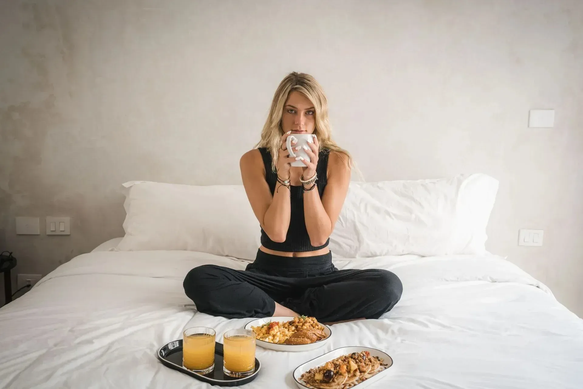 Woman sitting cross-legged on a bed, drinking from a mug, with breakfast tray.