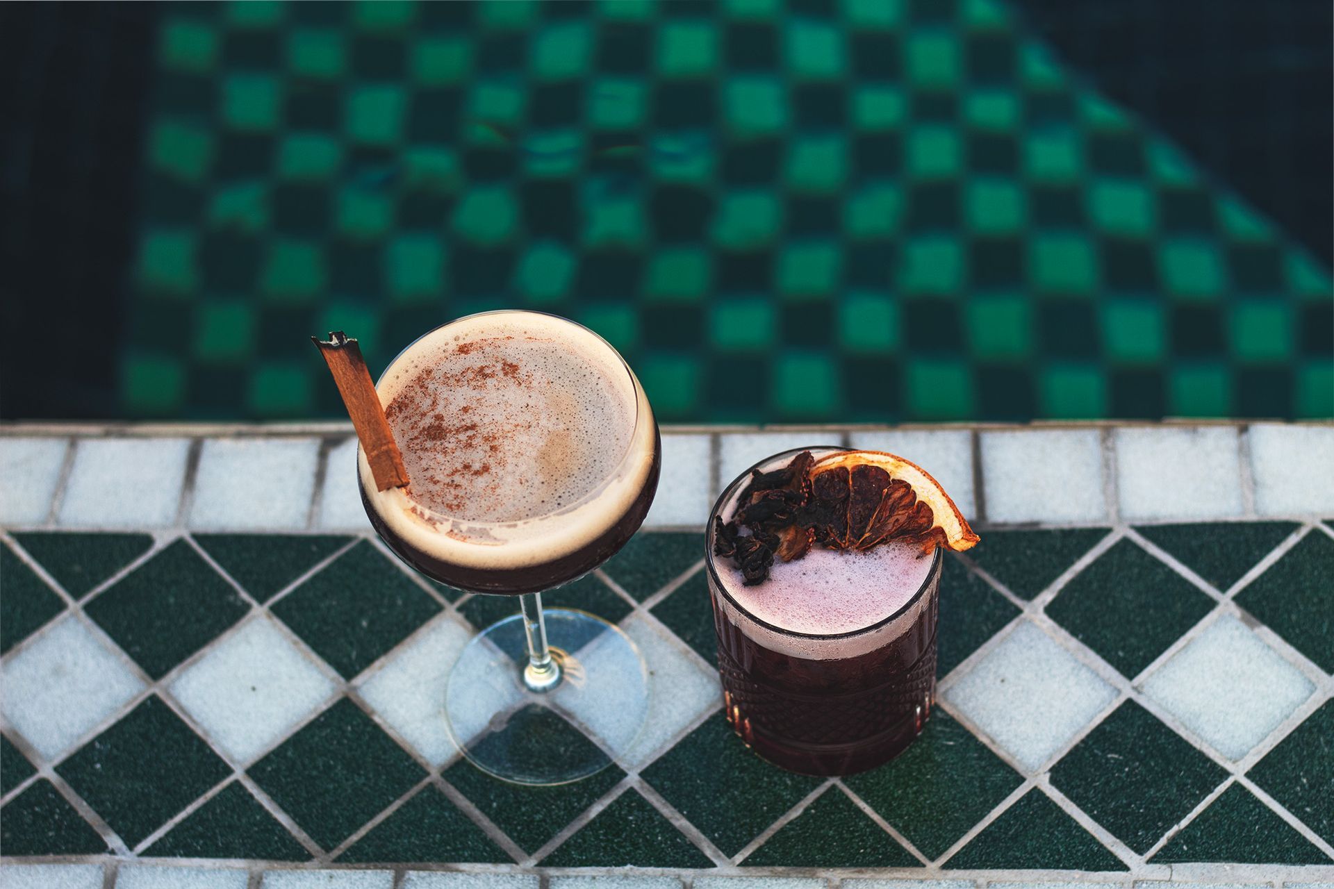 Hay dos bebidas sobre una mesa de azulejos al lado de una piscina.
