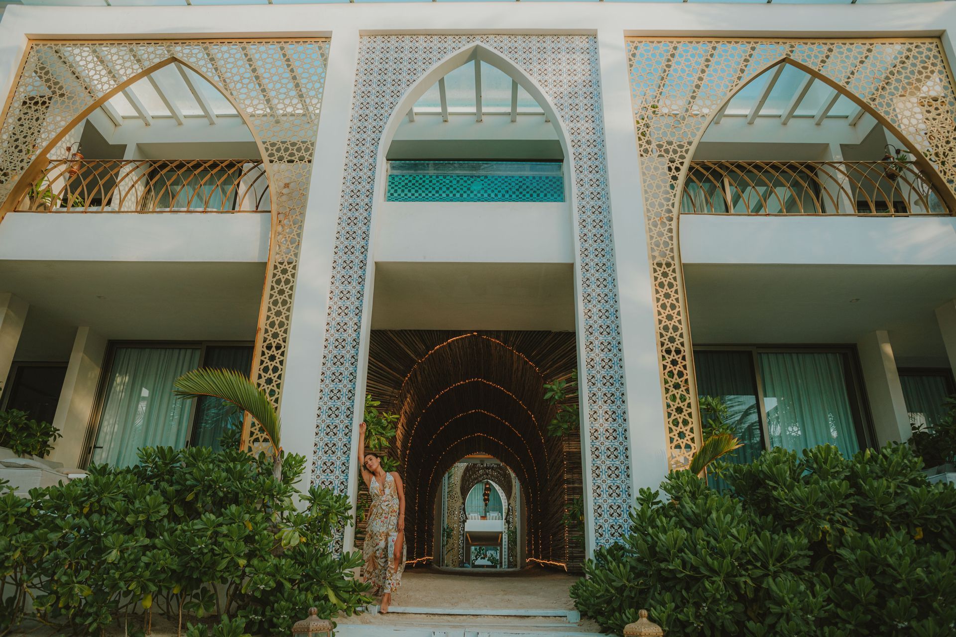 White hotel with arched entryways, decorative latticework, balconies, and lush green bushes.
