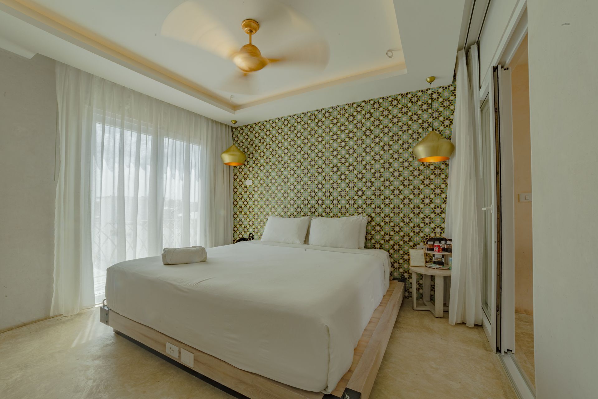 Dormitorio con una cama blanca, una pared con detalles estampados, apliques dorados y cortinas transparentes.