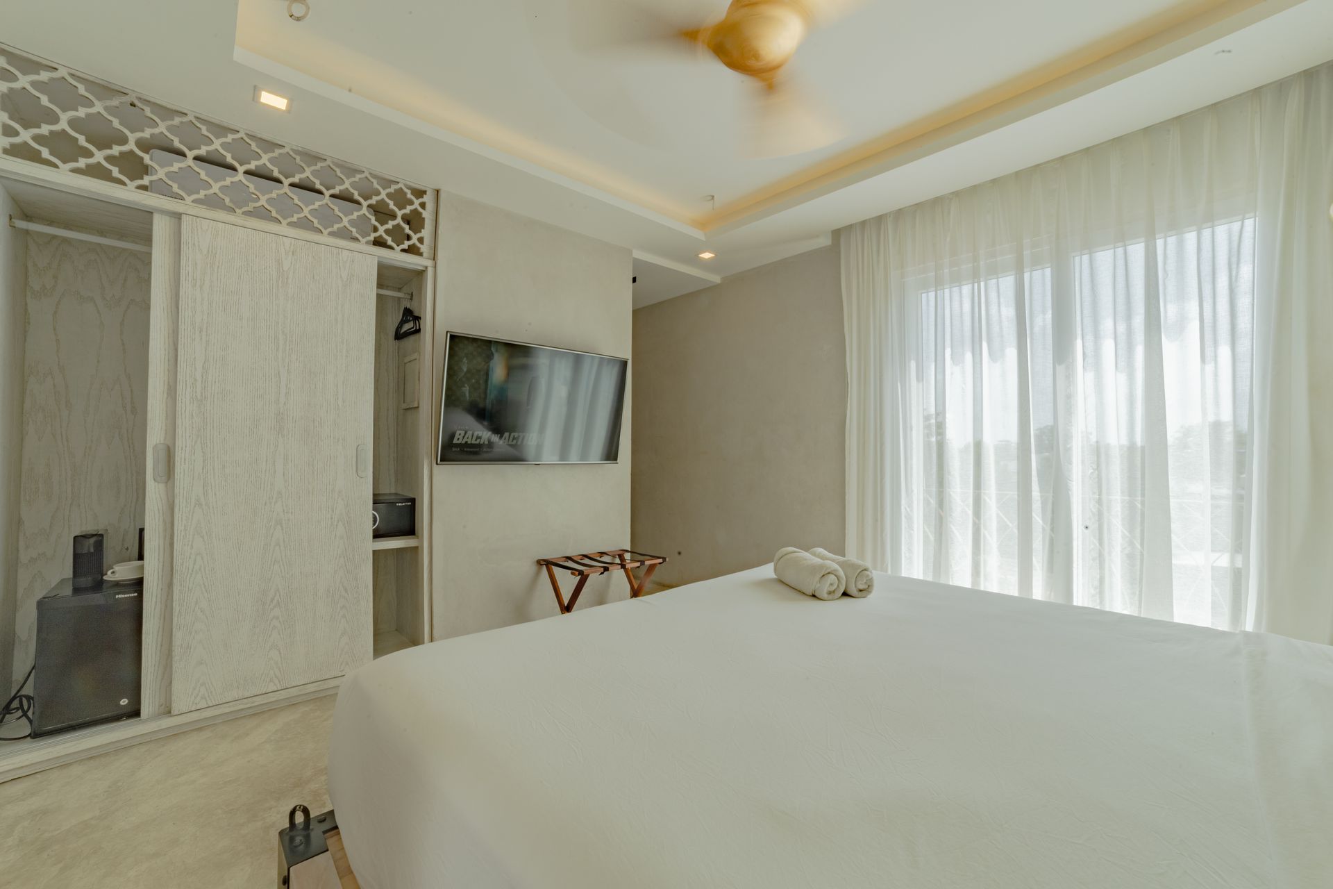 Dormitorio con cama blanca, armario y cortinas transparentes; televisión en la pared.