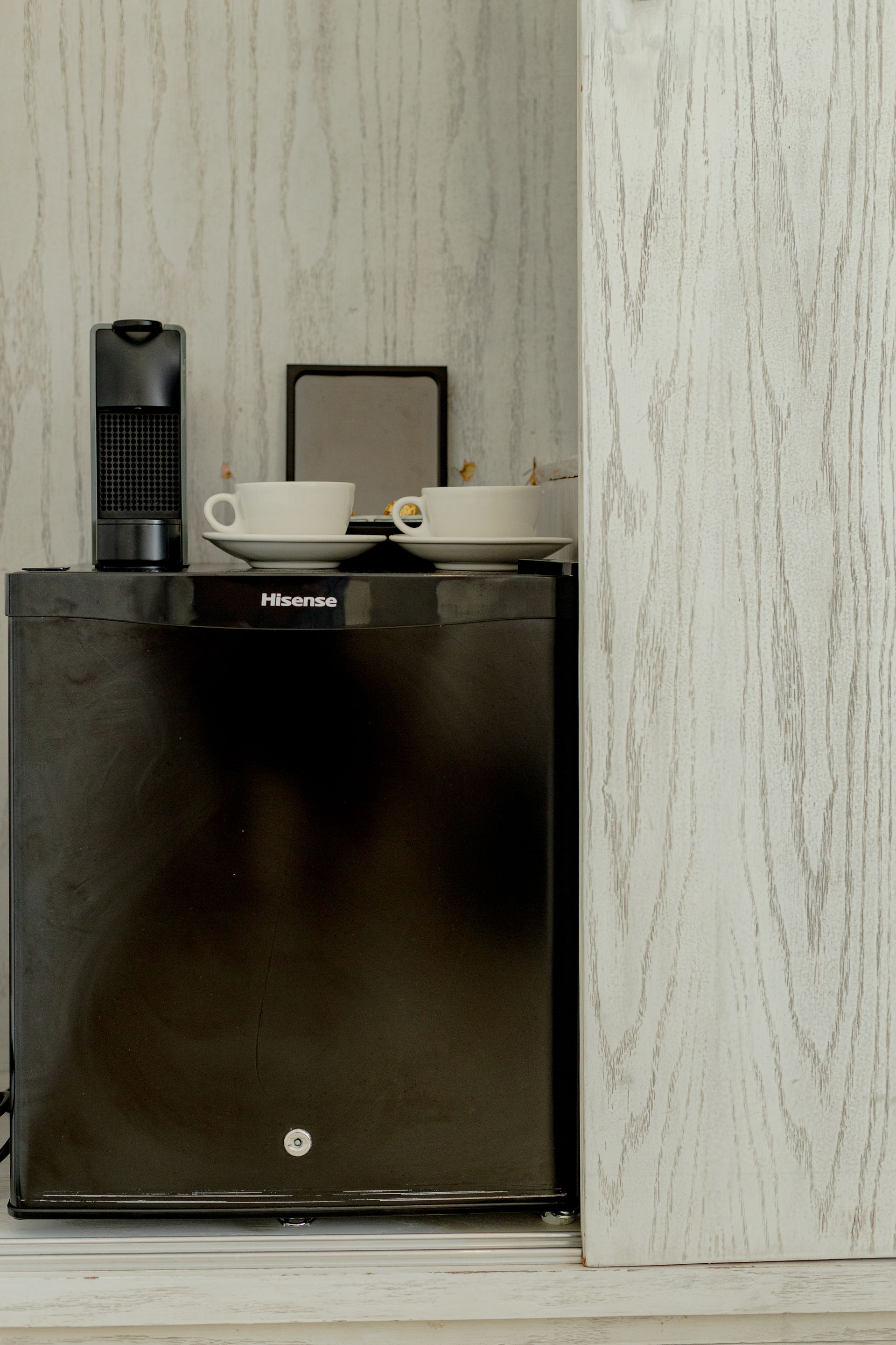 Mini-refrigerador negro con tazas de café en la parte superior, al lado de una pared con paneles de madera clara texturizada.