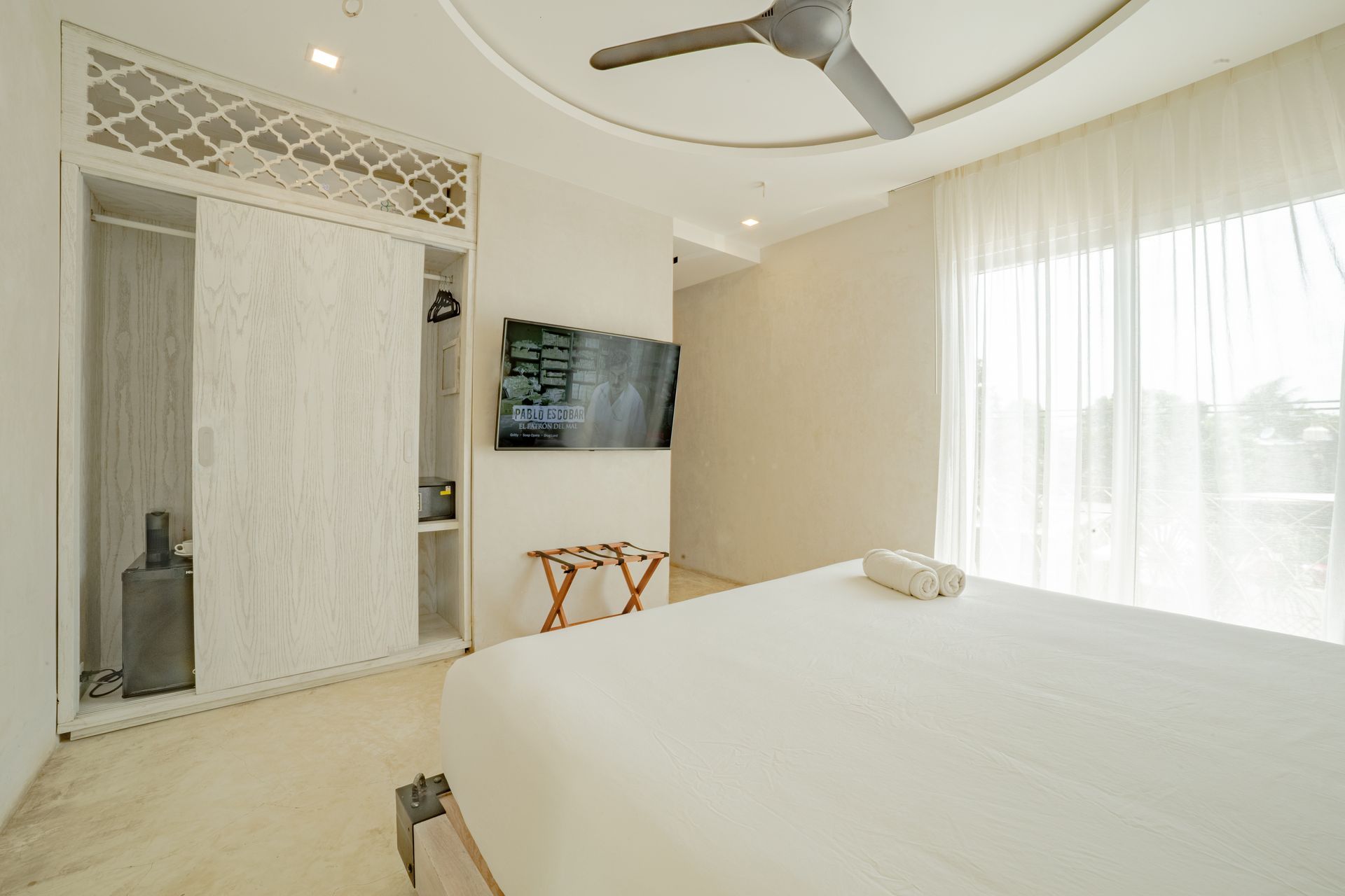 Dormitorio luminoso con cama blanca, armario y cortinas transparentes. Se ven un ventilador de techo y un televisor.