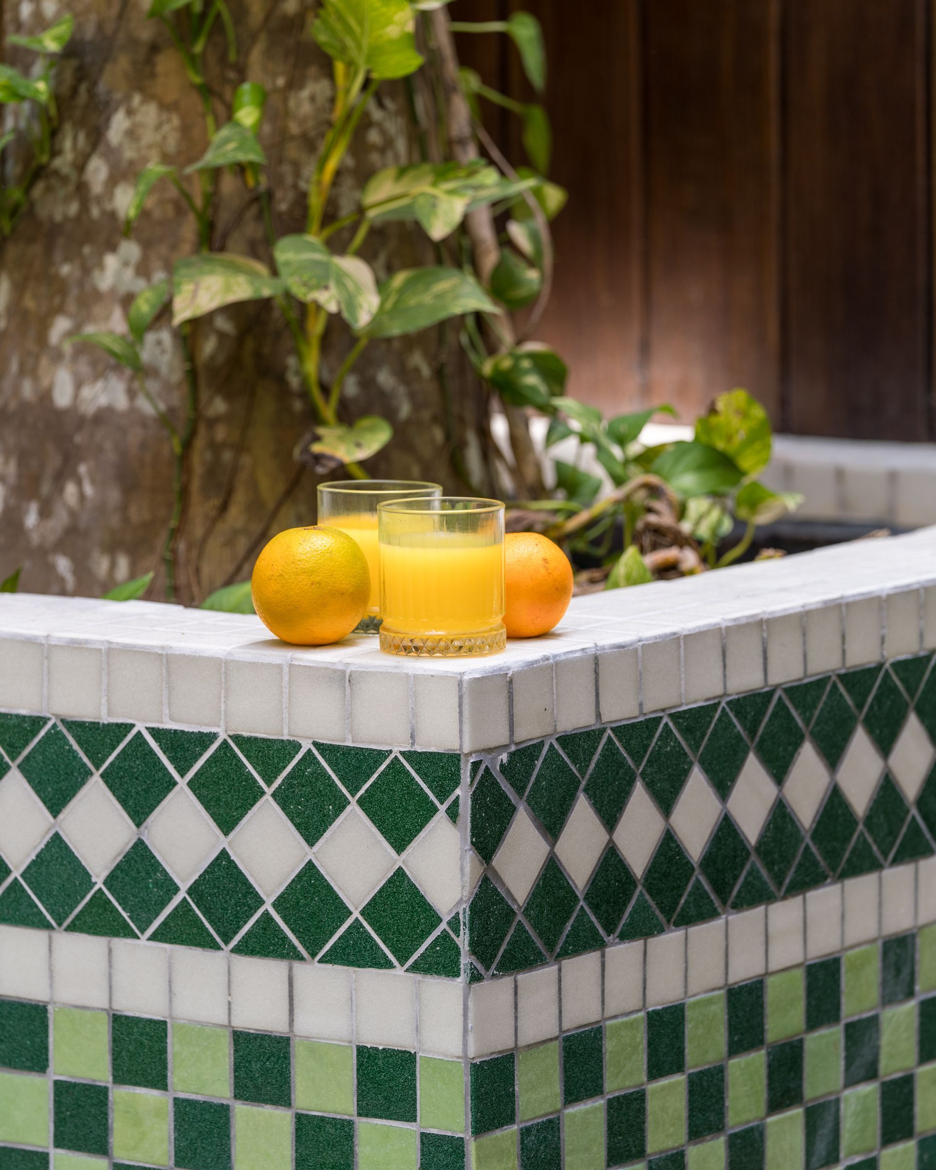 Two glasses of orange juice and two oranges are sitting on a tiled ledge.