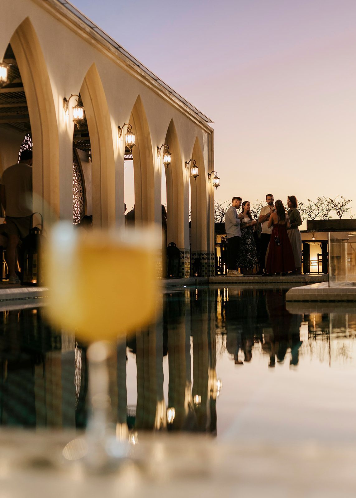 Un grupo de personas está de pie alrededor de una piscina con una copa de vino en primer plano.