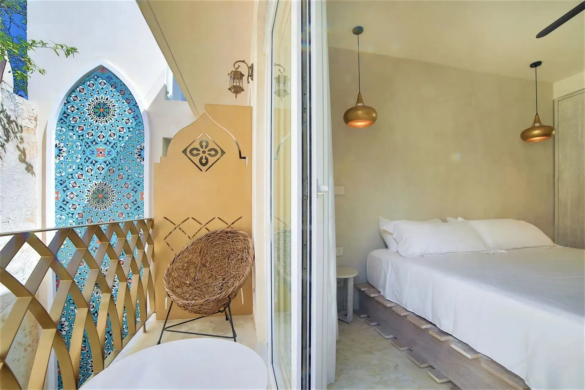 Dormitorio con balcón. Al abrir la puerta se ve una cama blanca, lámparas doradas, una silla de ratán y un mosaico turquesa.