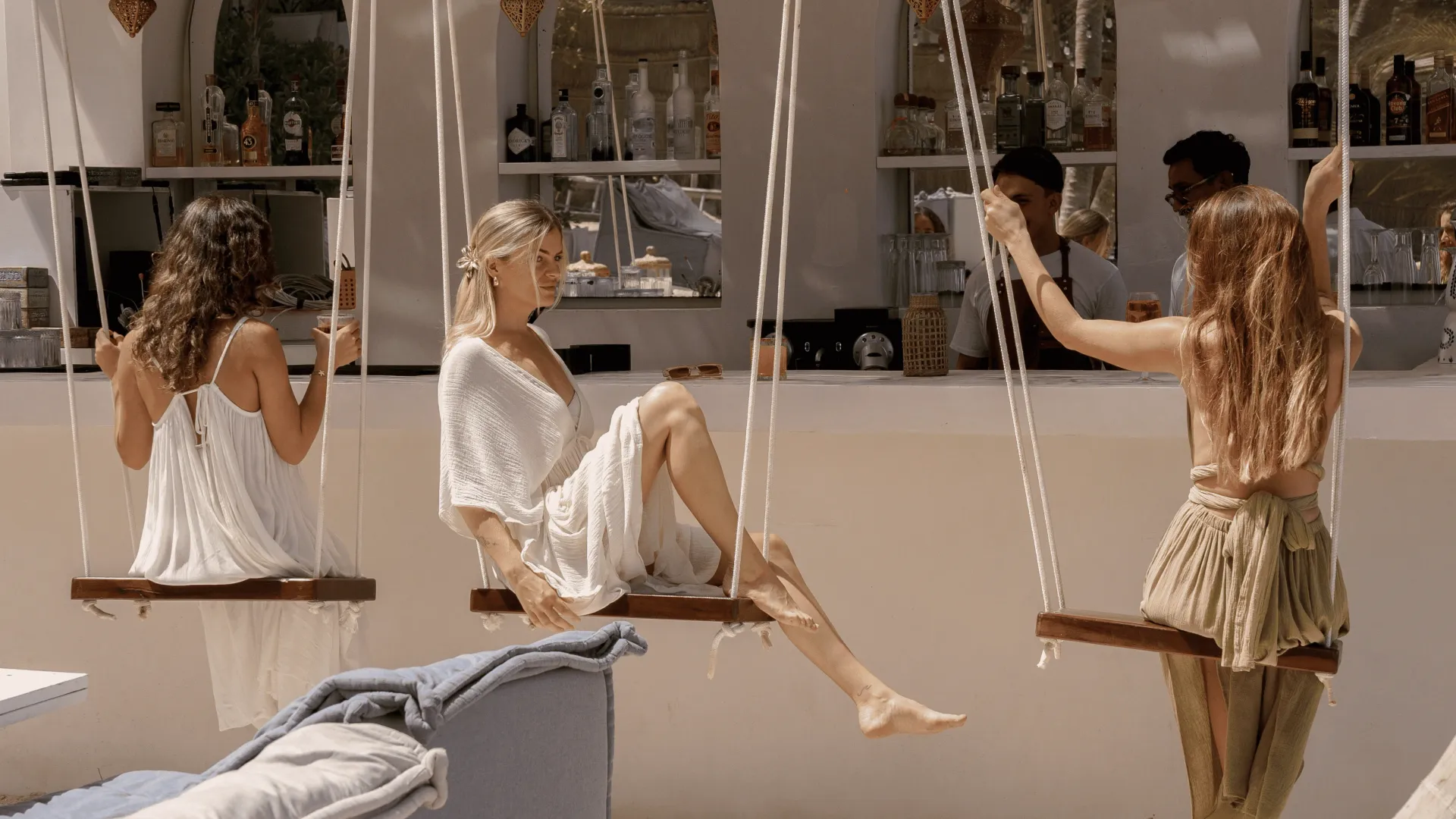 Three people on swings at a bar, sunlight. Drinks in background.