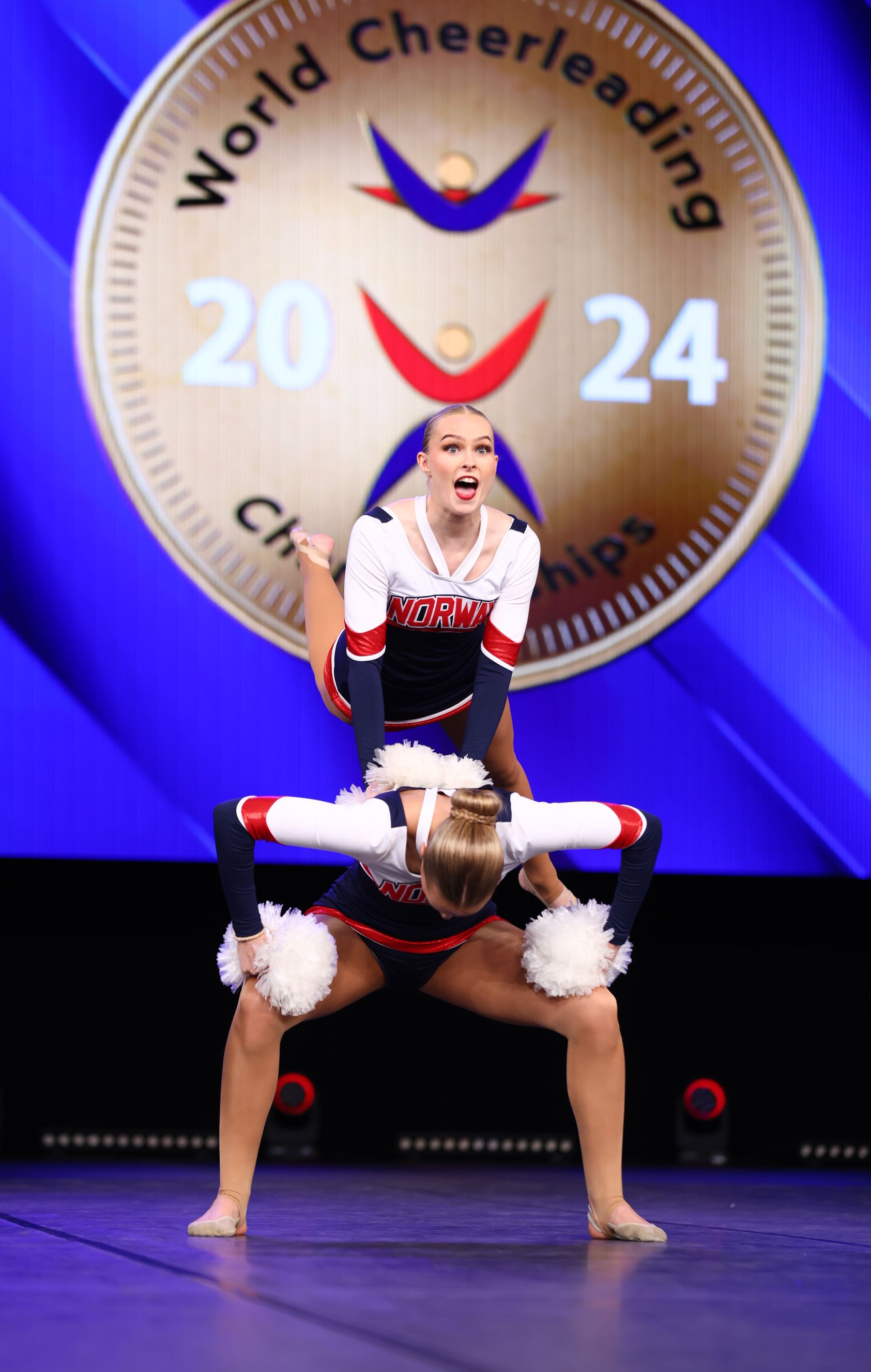 To cheerleaders i et stunt, mot en blå bakgrunn med en «World Cheerleading Championships 2024»-logo.
