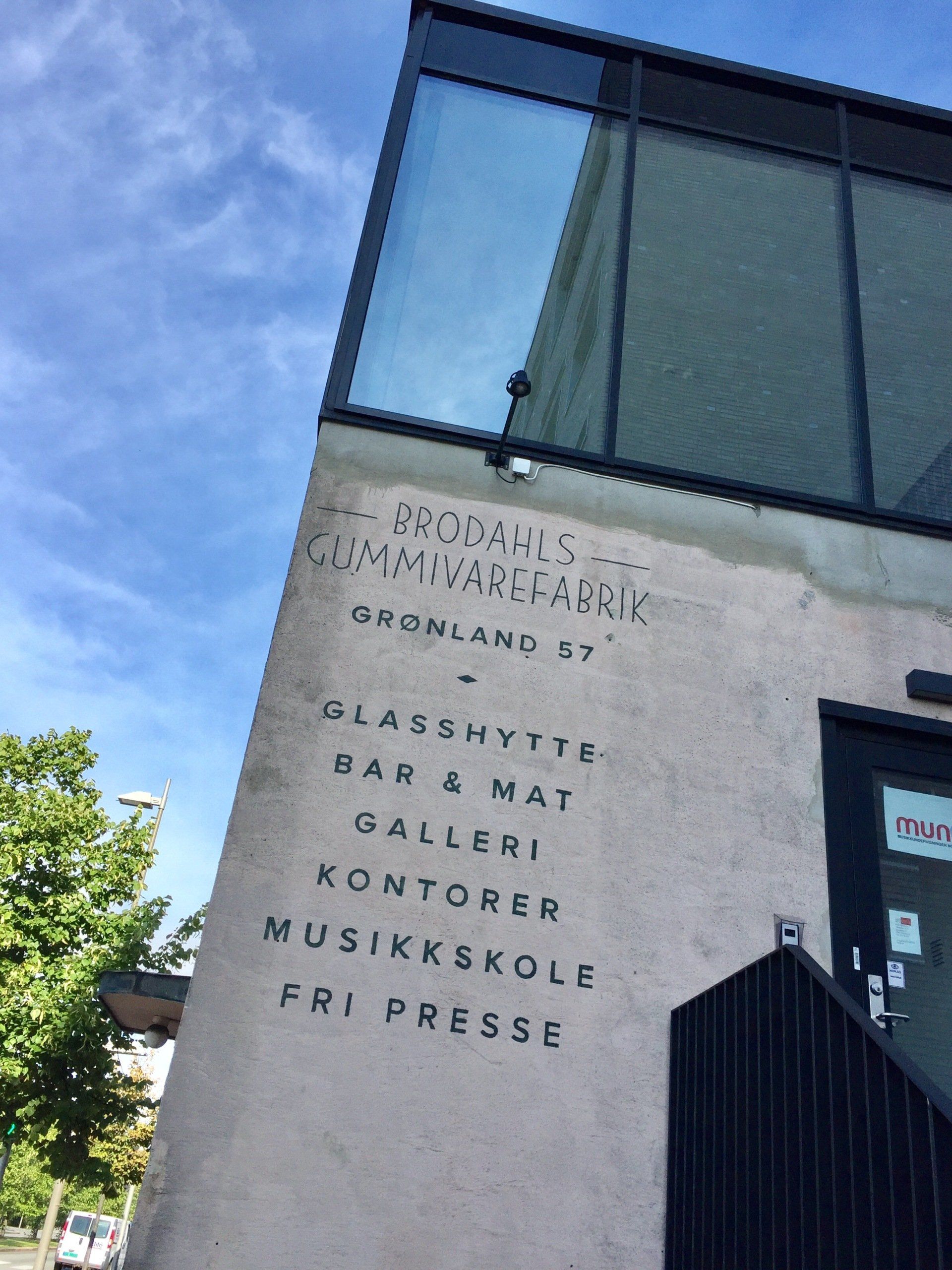 Vinklet bygning med tekst på siden; glassvinduer, en svart dør og blå himmel.