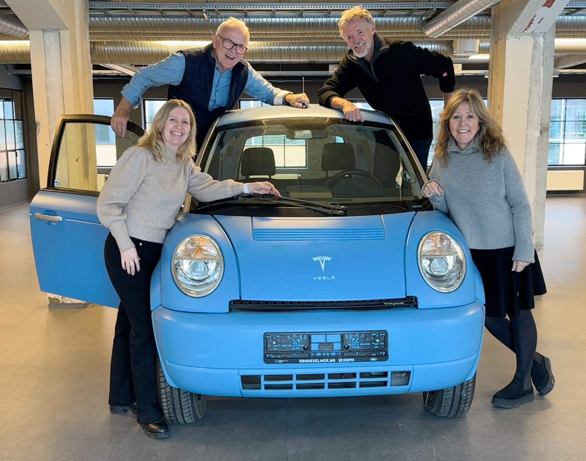 Four people pose with a light blue retro-style electric car indoors.