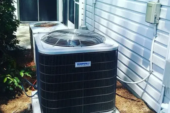 Outdoor air conditioning unit beside a house with white siding and plants nearby