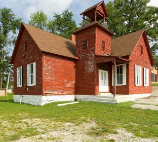 Red wooden schoolhouse with white shutters, a bell tower, and a small porch, standing in green grass.