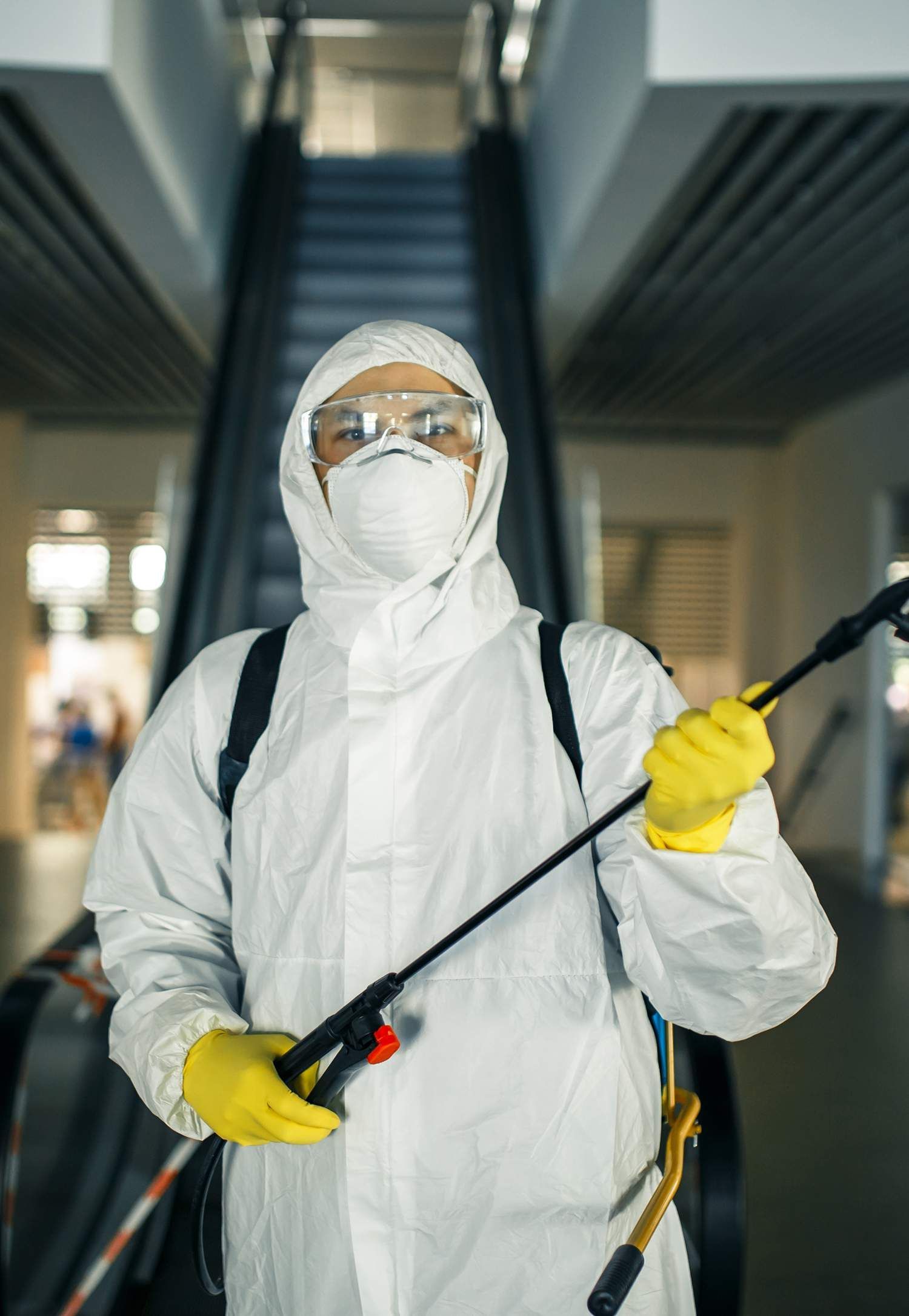 Person in hazmat suit spraying disinfectant near an escalator.