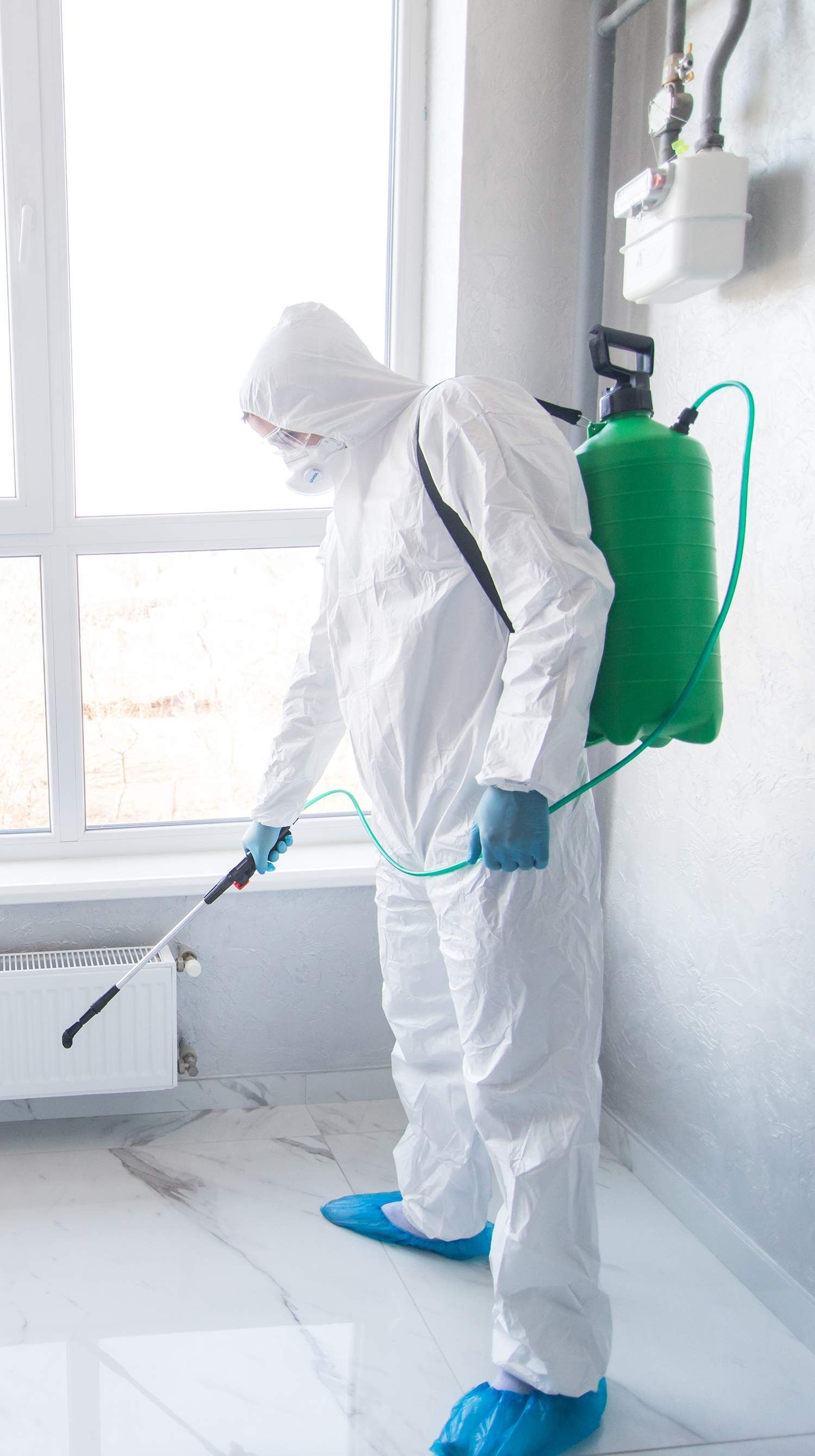Person in protective suit sprays disinfectant on a floor near a window.