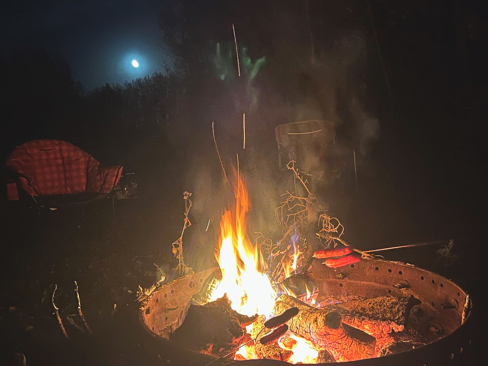 Campfire burning brightly at night with the moon in the background, hot dogs roasting.