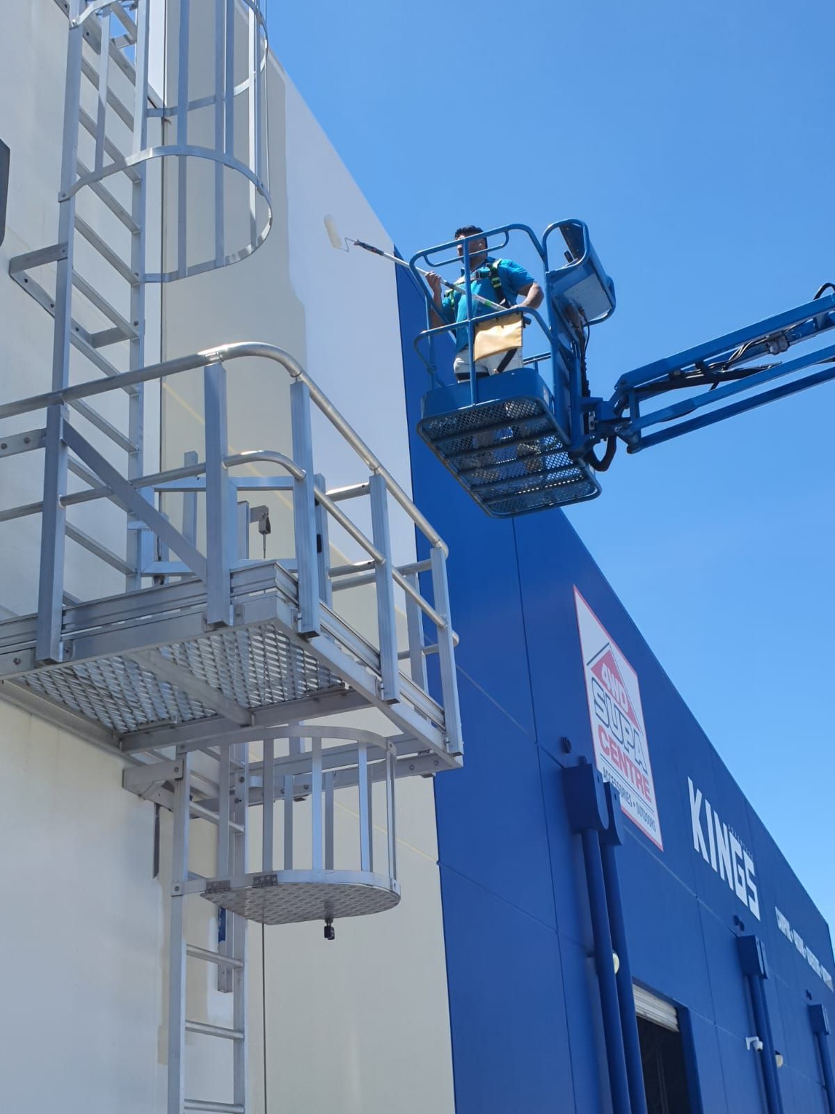 A person in a lift painting a blue wall. White ladder on the side. Clear blue sky.