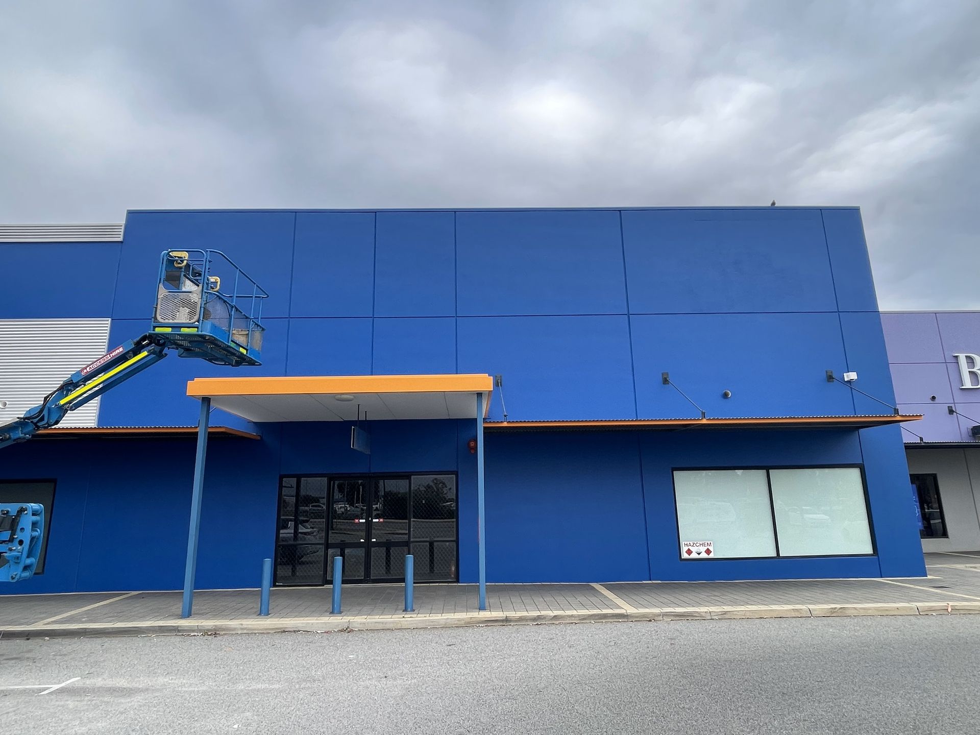 Exterior of a building being painted blue, with a cherry picker; cloudy sky.