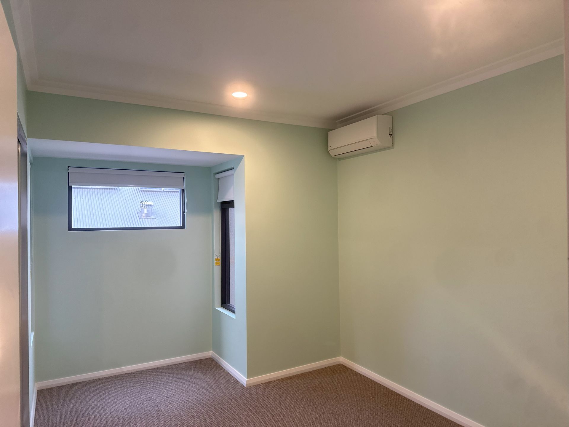 An empty room with light green walls, carpeted floor, and a window. A/C unit mounted on a wall.