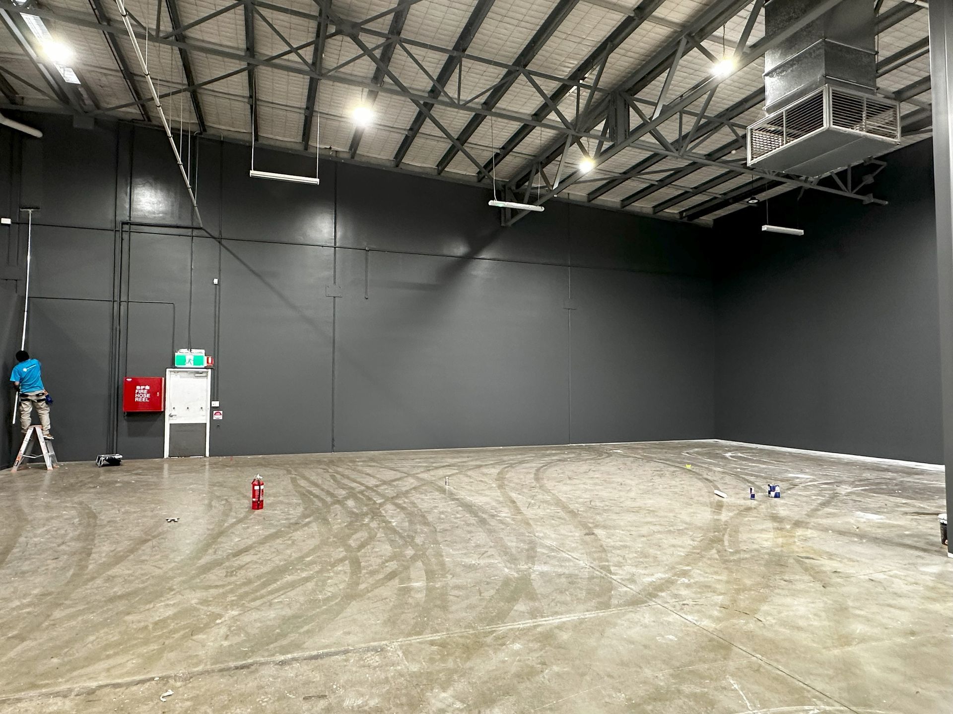 Empty gray warehouse interior with tire tracks on the floor, lit by overhead lights.