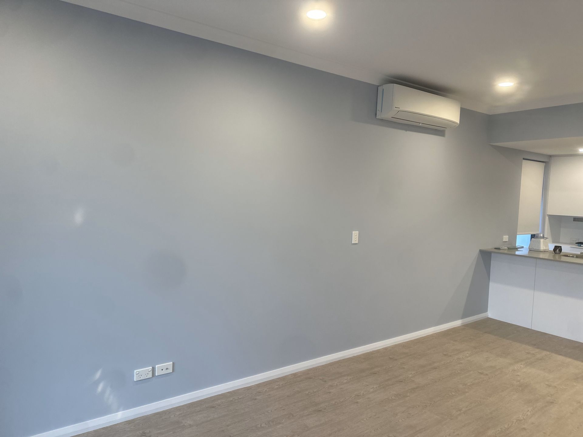 Empty room with light blue walls, air conditioner, and beige carpet. Kitchen area is visible.