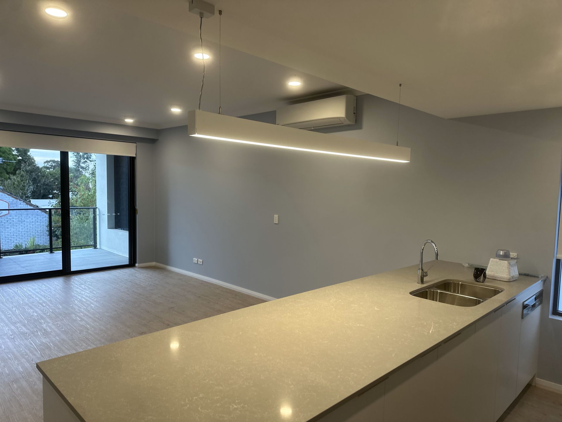 Modern kitchen and living area with light-colored walls and flooring, island with sink, and balcony access.