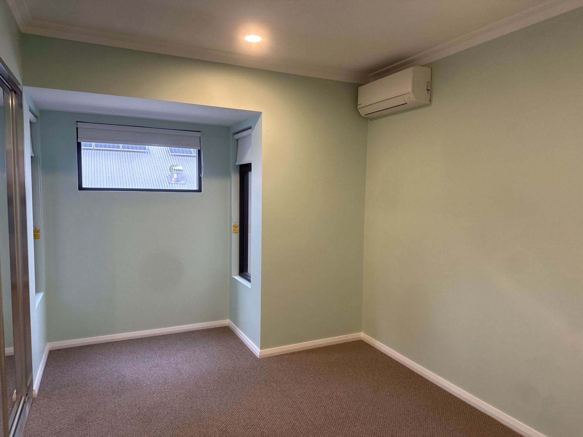 Empty room with light green walls, brown carpet, and an air conditioner. There are windows with a blind.