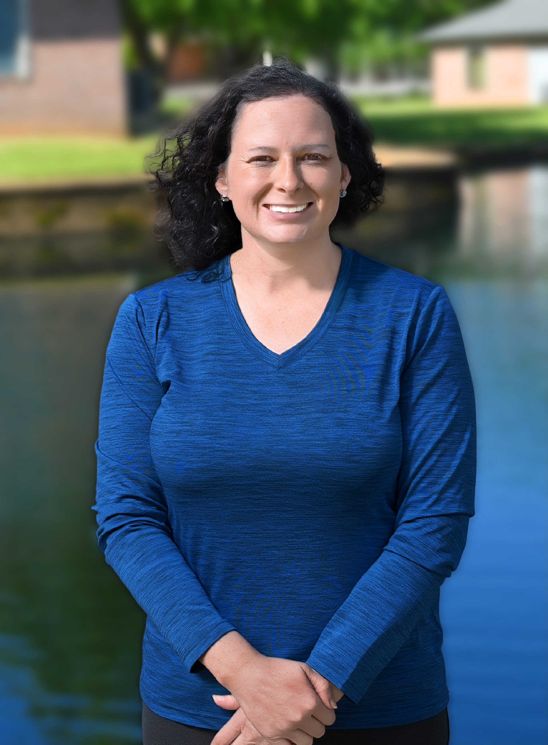 Smiling woman in a blue sweater standing outdoors by a pond, with blurred buildings and trees behind her