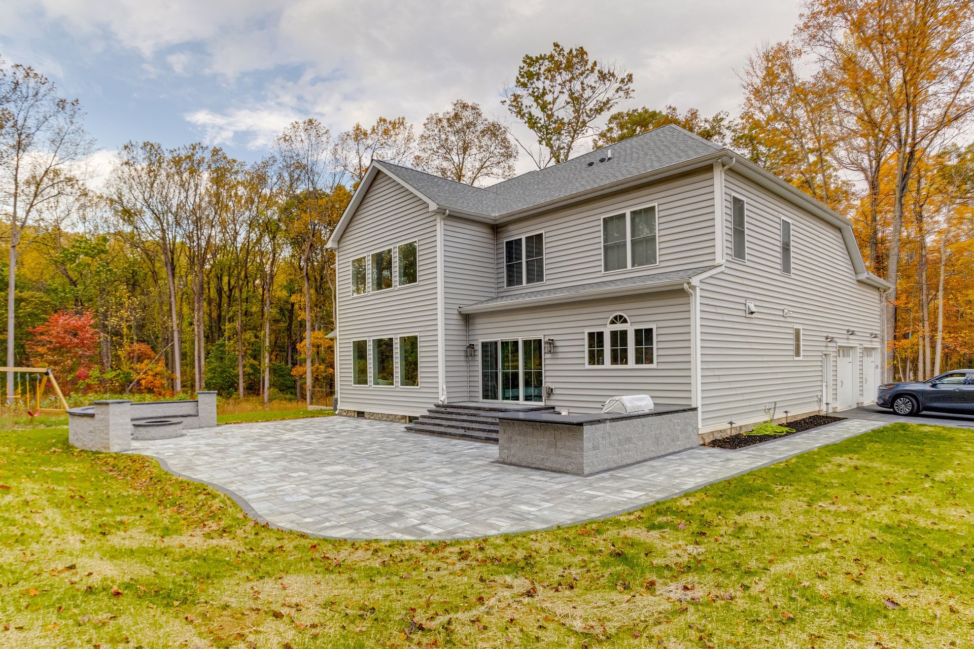 Back view of a gray two-story house with a stone patio and surrounding yard, set in a wooded area with fall foliage.