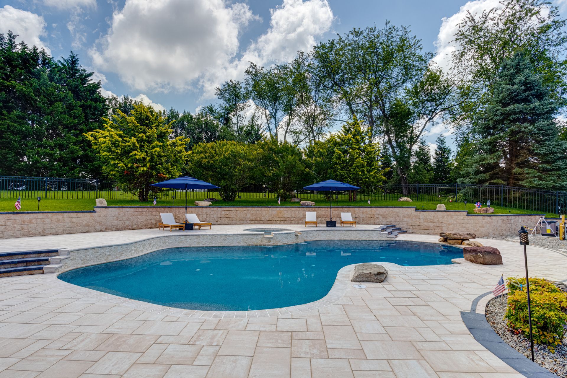 Swimming pool with lounge chairs, blue umbrellas, and lush greenery under a cloudy sky.