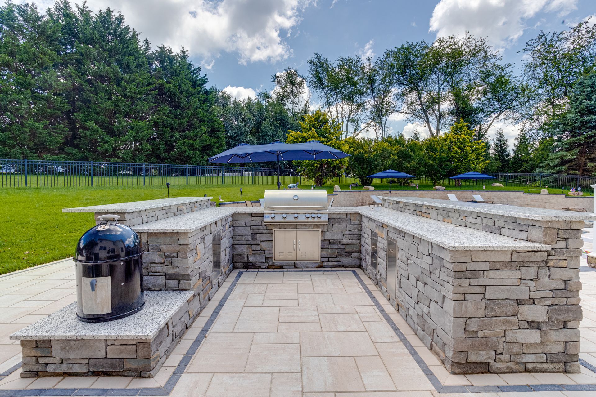 Outdoor stone kitchen with a granite-topped bar, built-in grill, and charcoal smoker on a stone patio in a grassy yard.