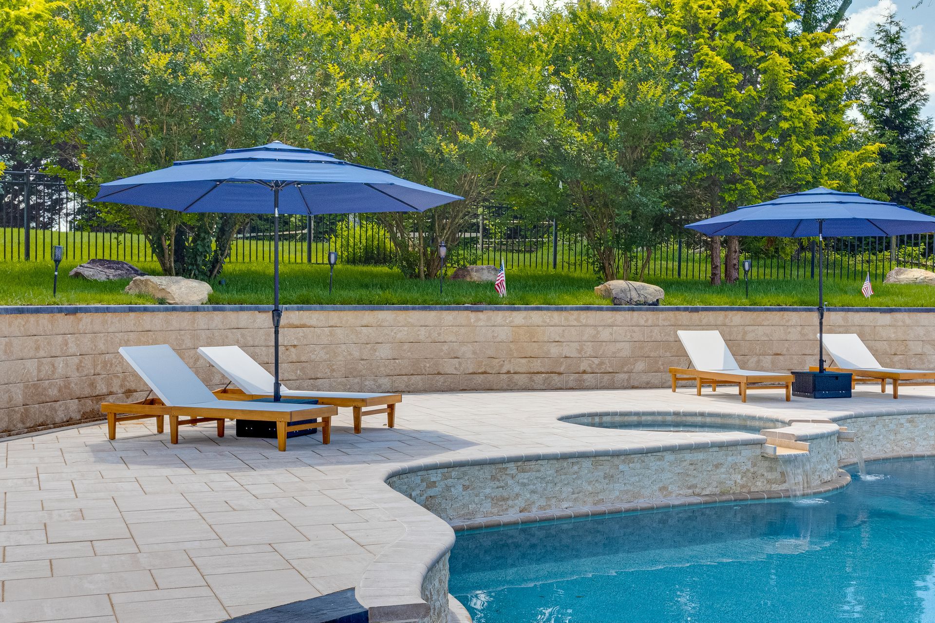 Poolside lounge chairs with blue umbrellas beside a curved swimming pool and stone pool deck
