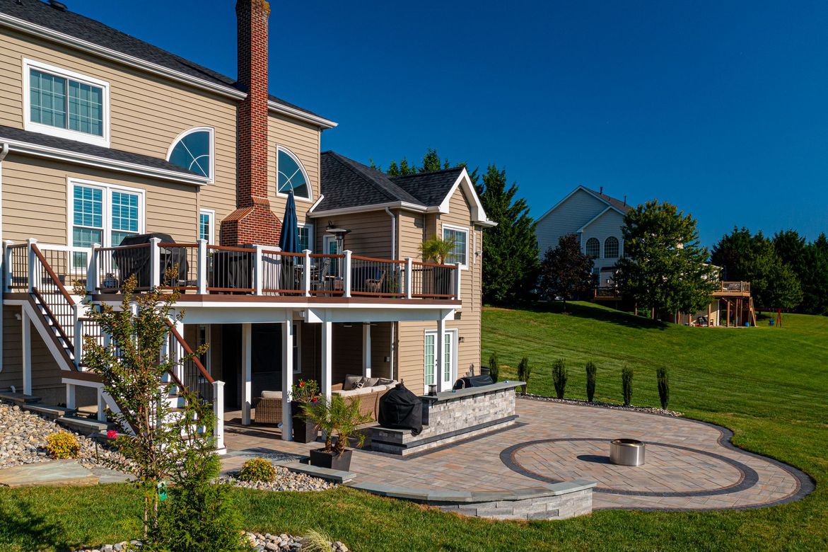 Two-story house with a deck overlooking a paved patio with a fire pit, set on a green grassy hill under a blue sky.
