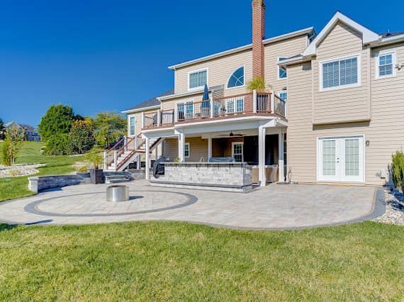 Backyard patio with a stone fire pit and outdoor kitchen, adjacent to a two-story beige house