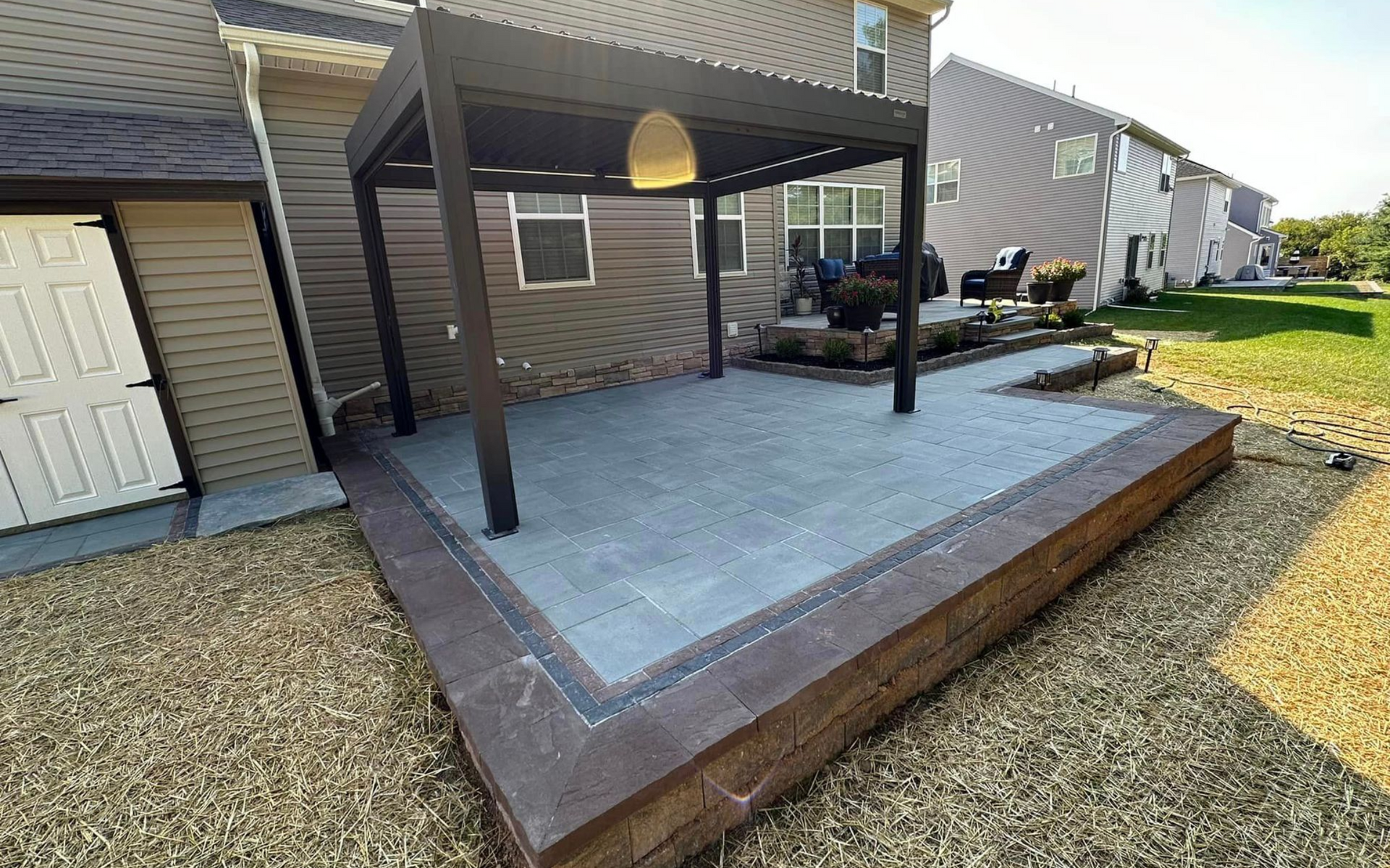 A patio with gray pavers and a dark wood pergola, enclosed by a stone retaining wall in a residential backyard.