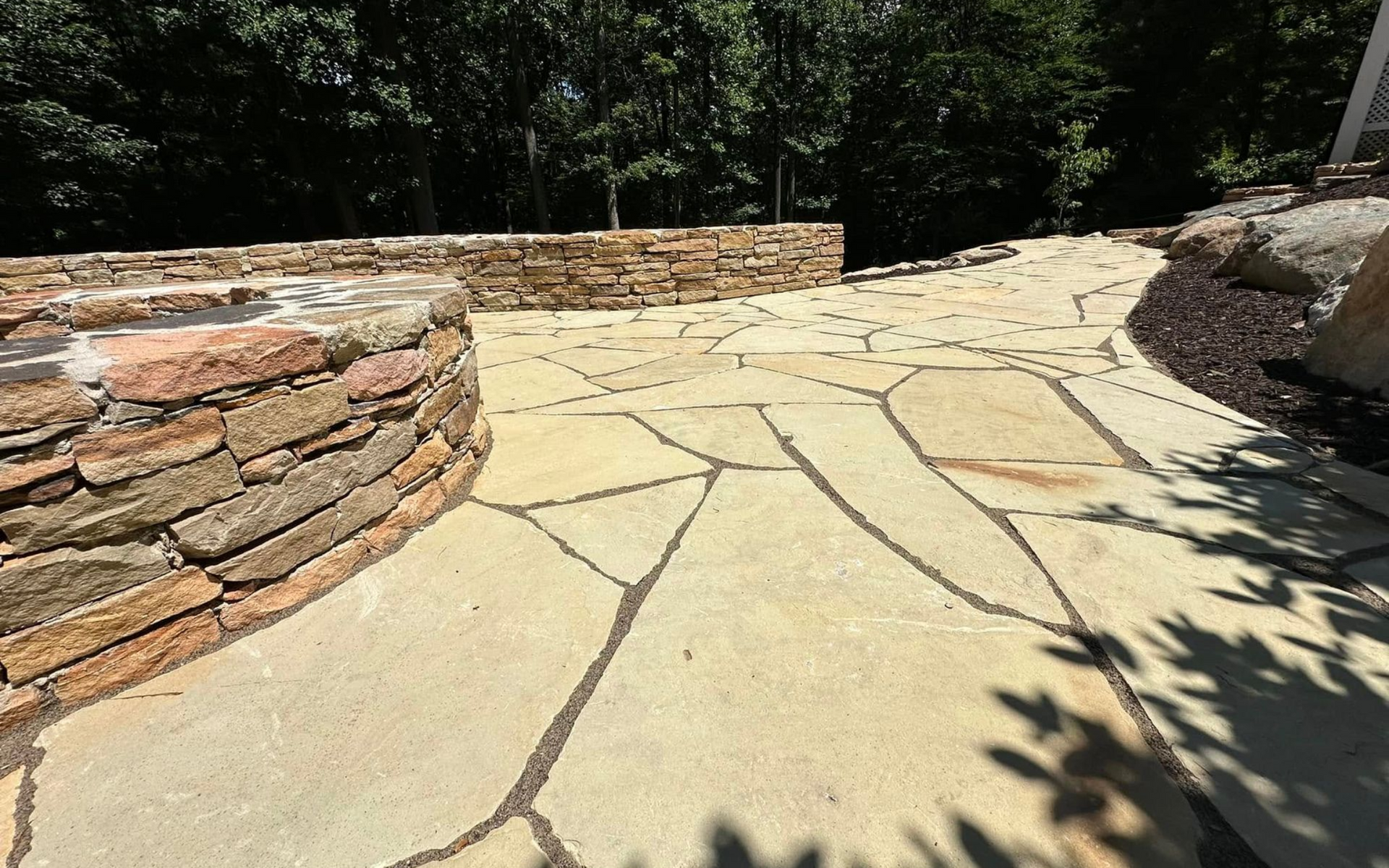 Flagstone pathway curving past a stone wall and into a wooded area. Sunny outdoor setting.