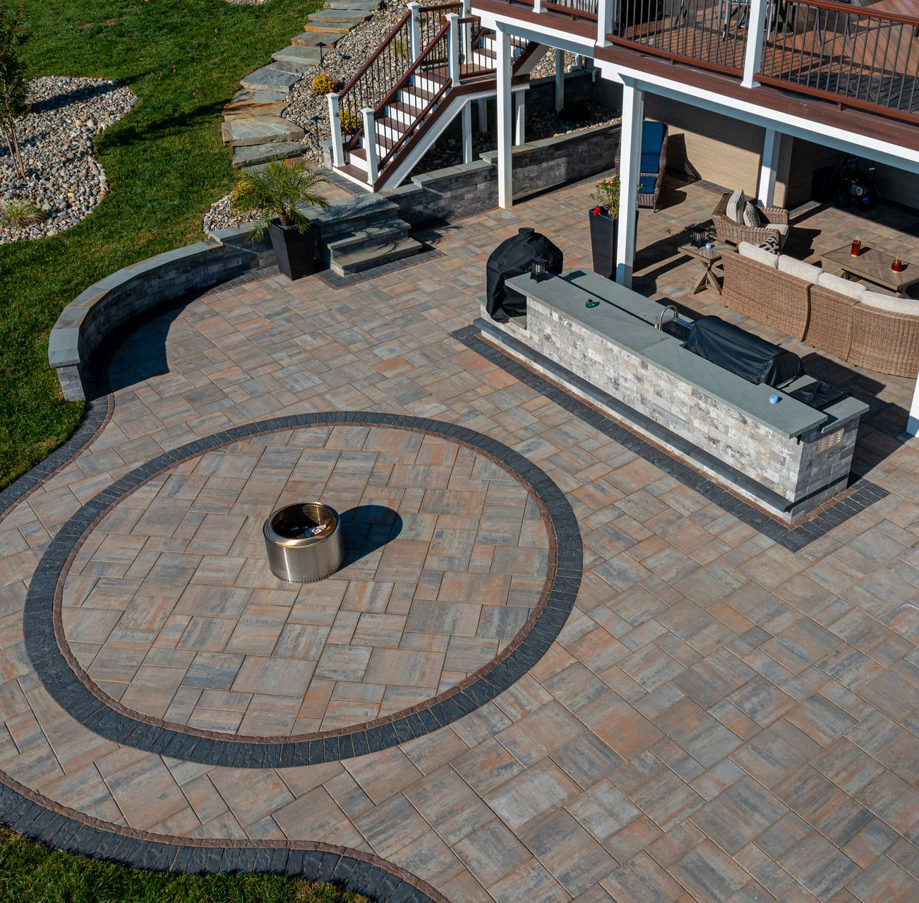Aerial view of a patio with fire pit, outdoor kitchen, and seating area. Brick pavers in neutral tones.